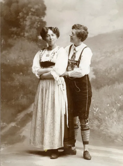 Two women holding around each other in a black and white photo, one is dressed in traditonal women's clothes, the other in men's clothes